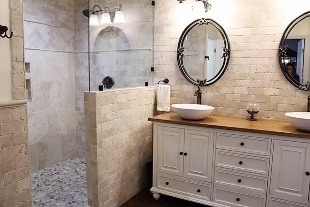 Bathroom with travertine tile walls, a pebble shower floor behind a tiled half wall and a white vanity with a vessel sink